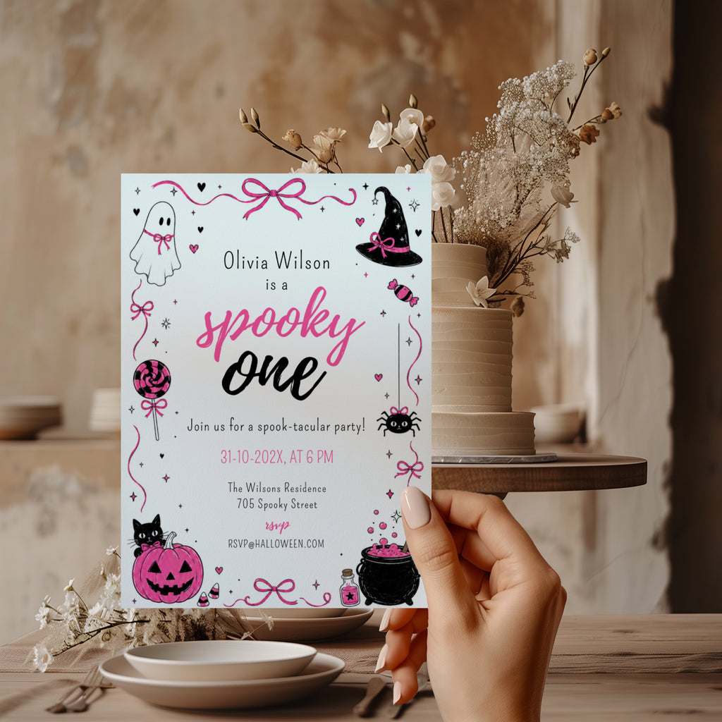 Spooky One first birthday girl invitation in pink and black with cute ghost, witch hat, spider, lollipop, pink pumpkin and bubbling Halloween cauldron illustrations.